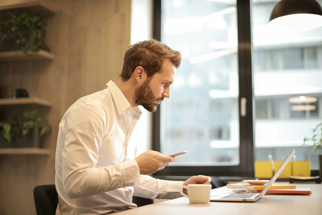 A man sits at his desk with his mobile phone in his hand while looking at his laptop. 