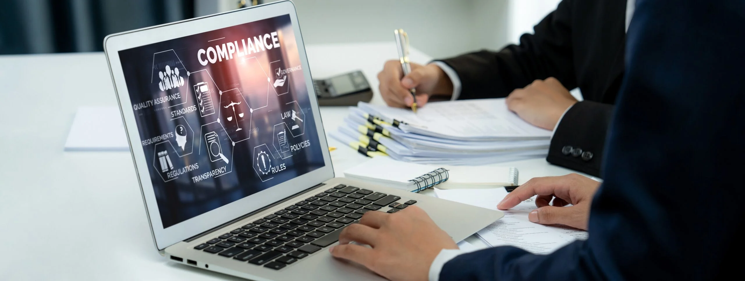 Business professionals reviewing compliance documentation while a laptop screen displays digital compliance, governance, regulations and policy icons.