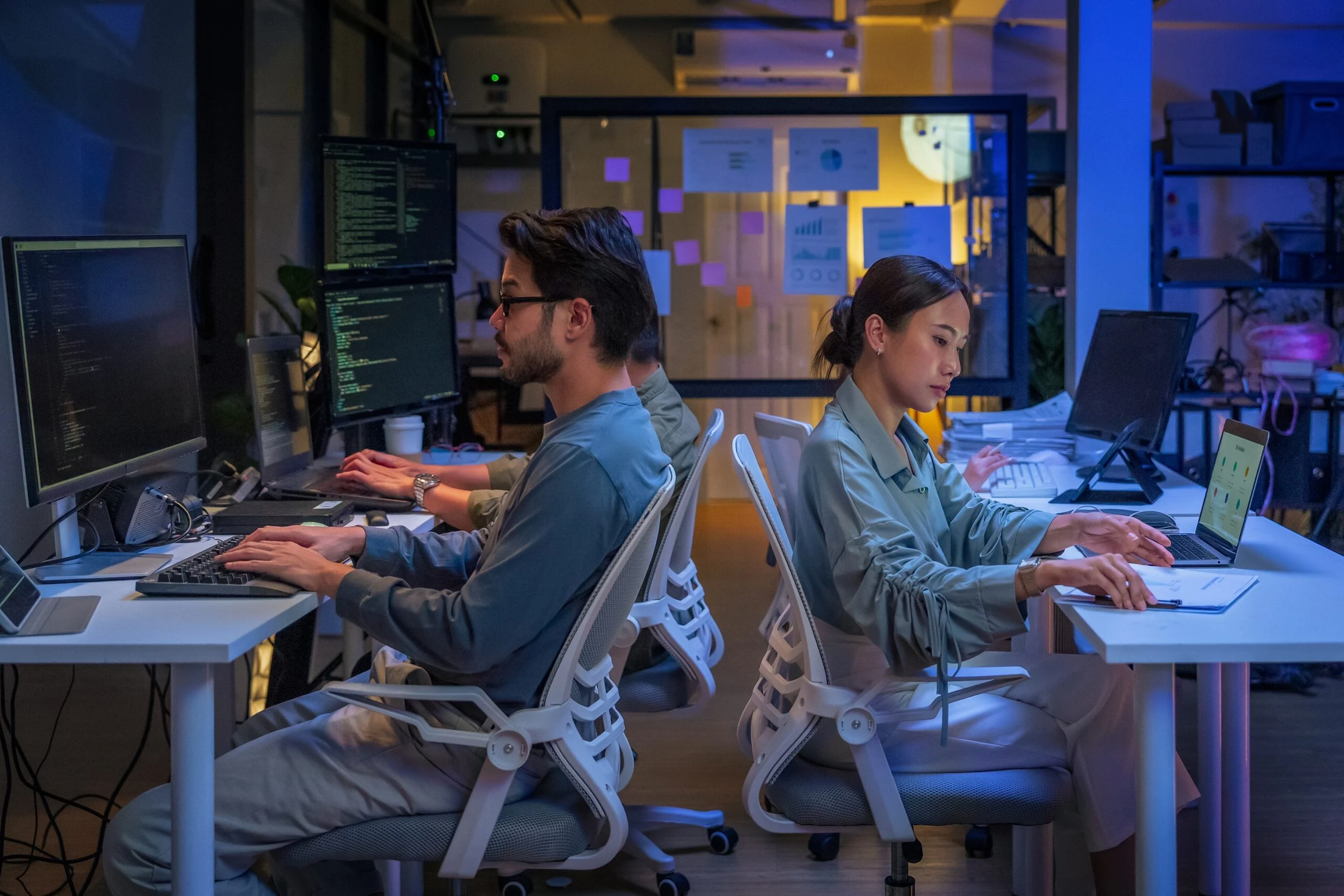 Two developers working at dual-monitor workstations in a modern office, reviewing code and data dashboards during a database migration for AI project.