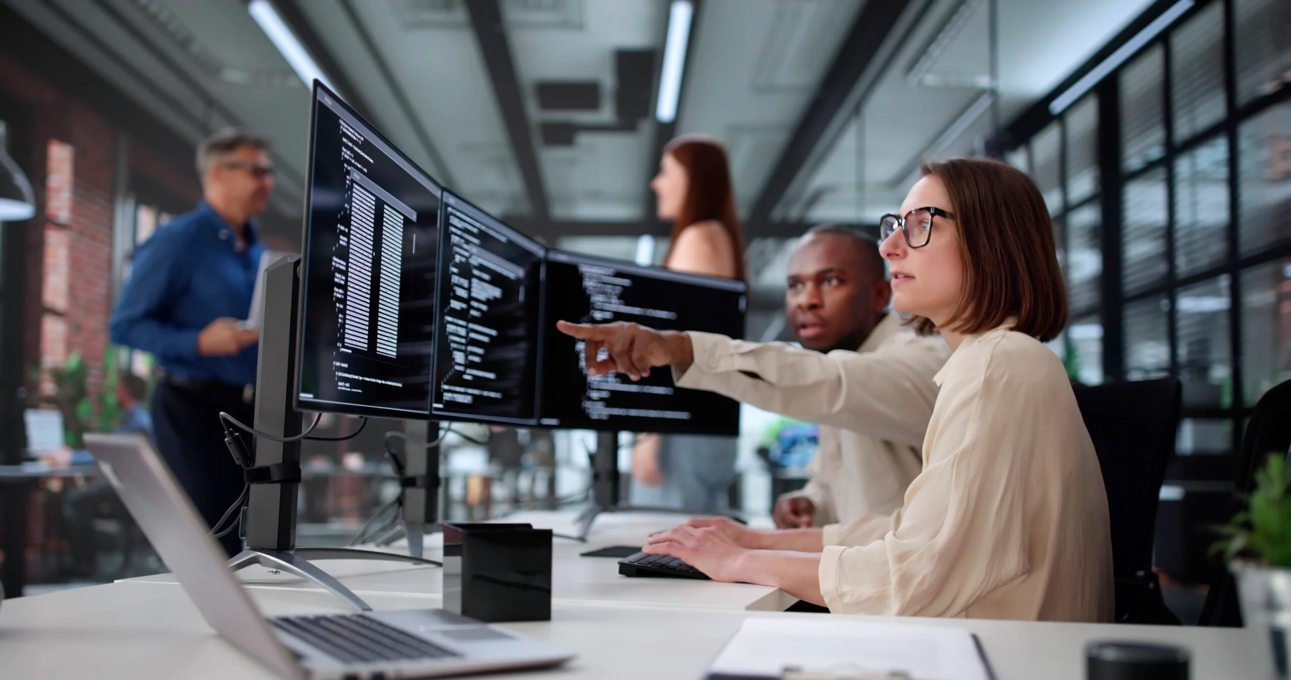 Two developers use a MongoDB AI assistant to review code and database queries across multiple monitors in a modern office